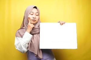Bastante joven mujer musulmana pensando, mano sosteniendo pancarta vacía en blanco, cartel, tablero blanco, tablero de letrero en blanco, tablero de anuncio blanco, presentando algo en el espacio de la copia, promoción