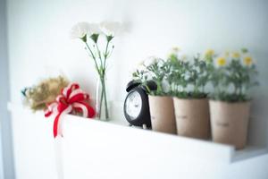 Vintage reloj despertador pone en el estante de madera blanca decorada por las flores en flor en la cerámica con fondo borroso de jarrón foto