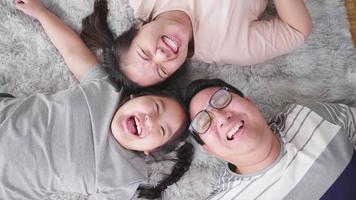 bovenaanzicht van gelukkige Aziatische familie liggend op de vloer in de woonkamer, camera kijken en samen lachen. in het weekend bij familie thuis blijven. gelukkig familieconcept video