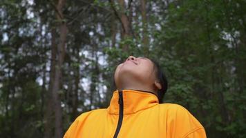 Happy Asian woman with black hair wearing yellow coat standing and taking a deep breath, getting some fresh air in the forest, traveling outdoor in beautiful nature. Trees background. Forest concept video