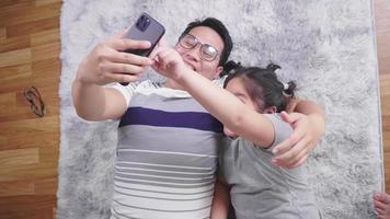 Top view of happy Asian dad and kid lying on the floor in living room and taking video call on smartphone. Staying with family at home in the weekend. Happy family concept