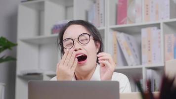 Asian woman getting sleepy during work, yawning and taking glasses off then taking it back and continue working on laptop, woking at home concept video