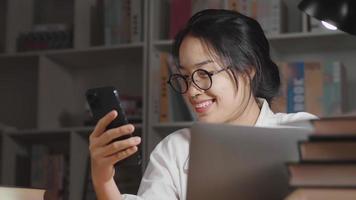 Happy Asian woman wearing glasses, using smartphone and looking at the laptop screen in working room at home at night. Taking relax from work. Working at home video
