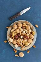 Mix of nuts on plate - walnut, almonds, pecans, macadamia. Healthy vegan food. Clean eating, balanced diet. Blue bright table, knife for opening shell. Top view, vertical photo