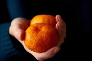 Close-up of woman's hand holding two whole bright citrus fruit tangerine on dark background.
