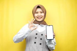 Hermosa joven musulmana asiática sonriendo confiada, entusiasta y alegre con la mano que sostiene el teléfono inteligente, promocionando algo, la aplicación de promoción de la mano, aislada sobre fondo amarillo