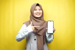 Hermosa joven musulmana asiática sonriendo confiada, entusiasta y alegre con la mano apuntando al teléfono inteligente, promocionando algo, aplicación de promoción manual, aislada sobre fondo amarillo foto