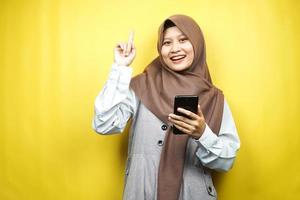Hermosa joven mujer musulmana asiática sonriendo, conmocionada, sorprendida, teniendo una idea, con las manos sosteniendo el teléfono inteligente, aislado sobre fondo amarillo, concepto de publicidad foto