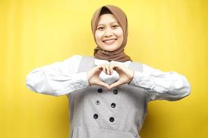 Hermosa joven musulmana asiática sonriendo confiada, entusiasta y alegre con las manos en señal de amor, afecto, feliz, en el pecho aislado sobre fondo amarillo foto