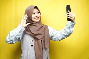Beautiful young asian muslim woman smiling confident, enthusiastic and cheerful with hands holding smartphone, calling someone, making video call, taking selfie, isolated on yellow background photo