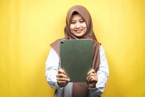 Beautiful young asian muslim woman smiling, excited and cheerful holding tablet, isolated on yellow background photo