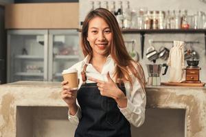 Close up of a young Asian female barista hold a cup of coffee serving to her customer with smile surrounded with bar counter background. Young female barista and her small shop. Food and drink concept photo