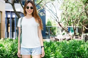 Asian hipster girl long brown hair in white blank t-shirt is standing in the middle of street. A female in street wear is standing on a green urban background. Empty mock up space for text or design. photo