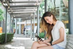 Portrait of attractive happy asian woman holding smartphone while sitting on roadside at the city street. Woman is sitting on the street near park. Summer holidays, road trip, vacation, travel concept photo