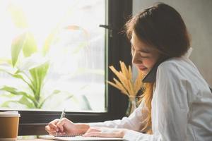 mujer de negocios joven en vestido blanco sentado a la mesa en la cafetería y escribiendo en el cuaderno. mujer asiática hablando teléfono inteligente y taza de café. autónomo que trabaja en cafetería. aprendizaje de los estudiantes en línea. foto