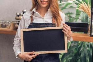 Young Asian female barista in denim apron hold a chalkboard with a beautiful smile in her own coffee shop welcome her customer to try a new coffee menu. Young female barista and her small coffee shop. photo