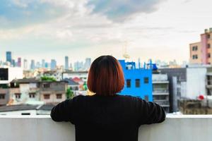 Alone asian woman sitting at rooftop of building see cityscape view in evening sunset time.lonely emotion photo