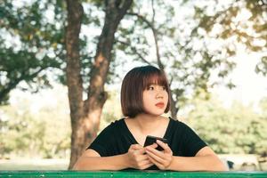Cute asian woman reading pleasant text message on mobile phone while sitting in park spring day. Asian woman using on smart phone with feeling relax and smiley face. Lifestyle and technology concepts. photo