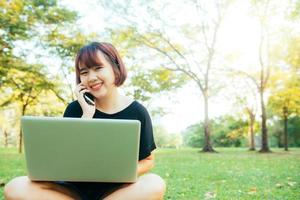 Cute asian woman smiles and talking on mobile phone while sitting in park spring day. Asian woman using on smart phone and laptop with feeling relax and smiley face. Lifestyle and technology concepts. photo