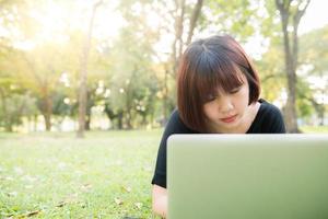 Young asian woman's legs on the green grass with open laptop. Girl's hands on keyboard. Distance learning concept. Happy hipster young asian woman working on laptop in park. Student studying outdoors. photo