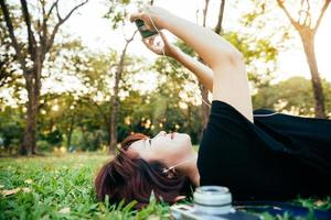 Young Asian woman laying on the green grass listening to music in the park with a chill emotion. Young woman relaxing on the grass with her camera beside. Outdoor activity in the park concept. photo