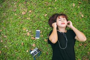 Young Asian woman laying on the green grass listening to music in the park with a chill emotion. Young woman relaxing on the grass with her music playlist. Outdoor activity in the park concept. photo