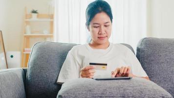 Mujer asiática que usa tableta, compra con tarjeta de crédito y compra Internet de comercio electrónico en la sala de estar de su casa cuando el distanciamiento social se queda en casa y el tiempo de auto cuarentena, compras en línea, concepto de coronavirus.