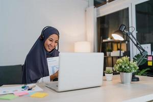 Asia muslim lady wear headphone using laptop talk to colleagues about sale report in conference video call while working from home office at night. Social distancing, quarantine for corona virus. photo