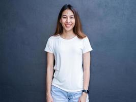 Asian hipster girl long brown hair in white blank t-shirt is standing in the middle of street. A female in street wear is standing on gray wall background. Empty mock up space for text or design.