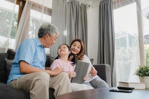 Asian grandparents and granddaughter using tablet at home. Senior Chinese, grandpa and grandma happy spend family time relax with young girl checking social media, lying on sofa in living room concept photo