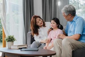 abuelos asiáticos hablando con su nieta en casa. Los chinos mayores, la generación anterior, el abuelo y la abuela que usan el tiempo en familia se relajan con la niña acostada en el sofá en el concepto de sala de estar. foto