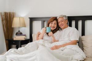 Asian senior couple talking on bed at home. Asian Senior Chinese grandparents, husband and wife happy drink coffee after wake up while lying on bed in bedroom at home in the morning concept. photo