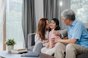 Asian grandparents kiss granddaughter cheek at home. Senior Chinese, old generation, grandfather and grandmother using family time relax with young girl kid lying on sofa in living room concept. photo