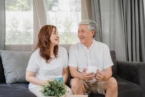 Asian senior couple relax at home. Asian Senior Chinese grandparents, husband and wife smile talking and drinking coffee while lying on sofa in living room at home concept. photo