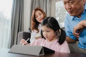 Asian grandparents and granddaughter video call at home. Senior Chinese, grandpa and grandma happy with girl using mobile phone video call talking with dad and mom lying in living room at home. photo