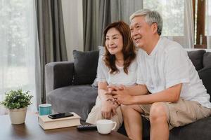 Asian elderly couple watching television in living room at home, sweet couple enjoy love moment while lying on the sofa when relaxed at home. Enjoying time lifestyle senior family at home concept. photo