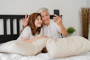 Pareja senior asiática selfie en casa. Abuelos chinos mayores asiáticos, marido y mujer felices usando selfie de teléfono móvil después de despertarse acostado en la cama en el dormitorio en casa en el concepto de la mañana. foto