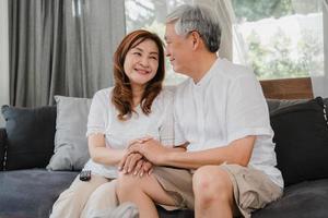 Asian senior couple relax at home. Asian Senior Chinese grandparents, husband and wife happy smile hug talking together while lying on sofa in living room at home concept. photo