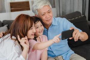 Asian grandparents selfie with granddaughter at home. Senior Chinese, grandpa and grandma happy spend family time relax using mobile phone with young girl kid lying on sofa in living room concept. photo