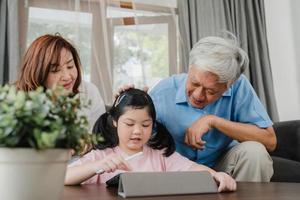 Asian grandparents and granddaughter video call at home. Senior Chinese, grandpa and grandma happy with girl using mobile phone video call talking with dad and mom lying in living room at home. photo