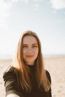 Retrato de mujer bonita con cabello largo, rubia toma selfie en teléfono móvil en la playa de arena foto
