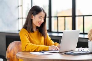 Asian woman working on laptop at home or in cafe. Young lady in bright yellow jumper photo