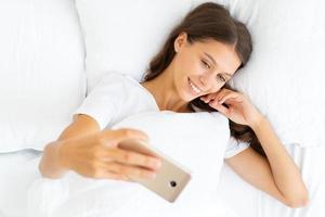 Bonita selfie femenina, rubia toma una foto en el teléfono móvil en la cama. hermosa mujer mirando a la cámara