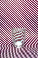 A glass of water with a double bottom on a striped background. Abstract refraction of red and white diagonals in glass. Vertical format photo