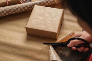 Cerrar mujer mano cortando papel kraft para envolver la caja de regalo de navidad en la mesa de madera