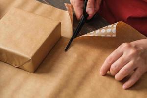 Cerrar mujer mano cortando papel kraft para envolver la caja de regalo de navidad en la mesa de madera
