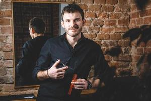 Portrait male barber holds sharp scissors and comb against a brick wall background photo