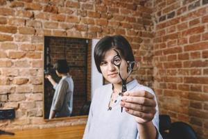Young beautiful hairdresser woman with scissors in hand against a brick wall background