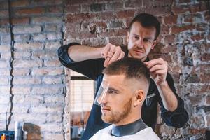 joven barbero afeita la cabeza del cliente con una recortadora eléctrica. Joven barbudo cortarse el pelo por peluquero en peluquería foto