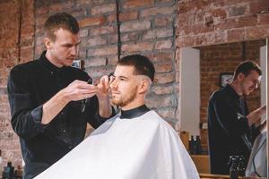 Macho joven barbero afeita la cabeza del cliente con una recortadora eléctrica foto